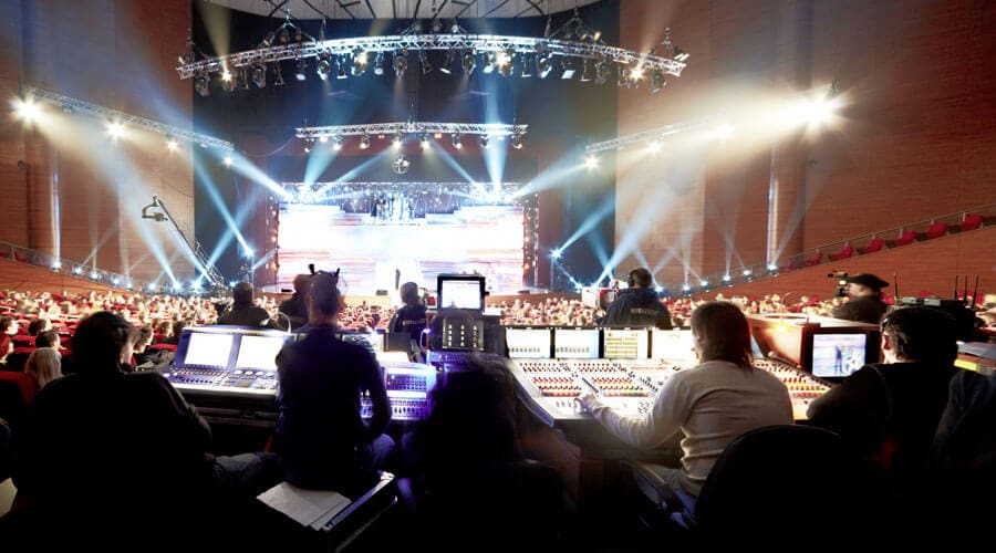 A live event is supervised by a team of technicians in the control booth, where lighting and sound are set up for the performance. Bright stage lighting and various technical devices are used to enhance the overall experience for the audience.