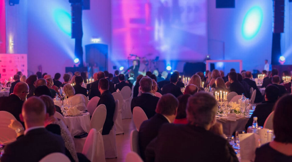 A large group of elegantly dressed people sit at round tables and take part in a formal event. Soft, colorful lighting and candles create a warm and inviting atmosphere in the room.