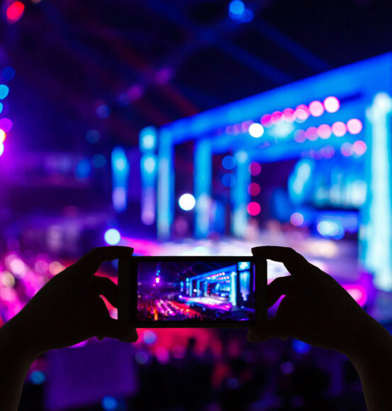 Im Vordergrund hält eine Person ein Smartphone in der Hand und nimmt das Geschehen über den Bildschirm des Geräts auf, auf dem deutlich die beleuchtete Bühne zu sehen ist, oder fotografiert es. Der Hintergrund ist mit unscharfen, aber hellen Lichtern gefüllt, was darauf hindeutet, dass durch die Lichteffekte auf der Bühne eine energiegeladene Atmosphäre geschaffen wird