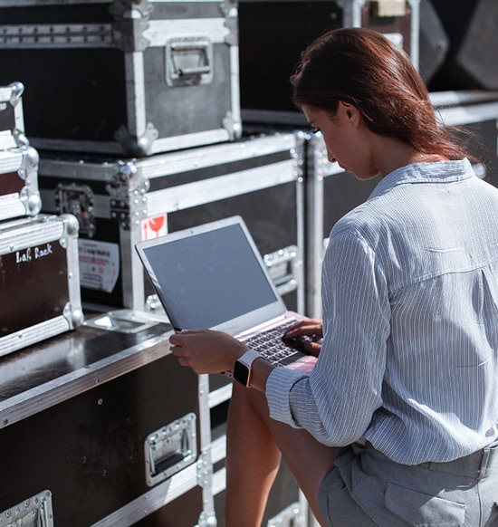 A young woman with brown hair and a light grey shirt is sitting in front of cases holding a laptop on her knees.