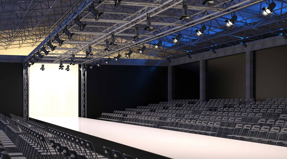 An empty runway is being prepared for a fashion show, with rows of chairs arranged on either side for the audience. Overhead lighting and staging equipment are being set up to illuminate the catwalk area for the upcoming event.