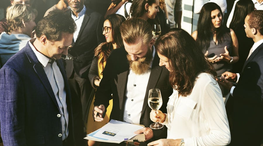 A group of people is being shown at a business event, where documents are being reviewed and conversations are being conducted. Drinks are being held by the participants as they interact with one another.