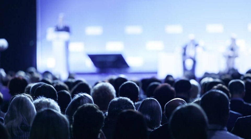 Ein Publikum ist in einem Konferenzsaal versammelt und hört aufmerksam den Rednern zu, die auf der Bühne stehen. Die Veranstaltung wird mit sanftem blauen Licht beleuchtet, das eine professionelle und konzentrierte Atmosphäre schafft.
