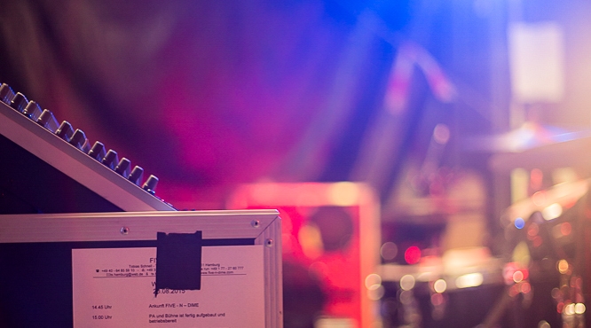 A part of a mixing console is being shown in the foreground with a taped setlist attached to it. In the background, a blurred scene with colorful lights and musical instruments is being depicted.