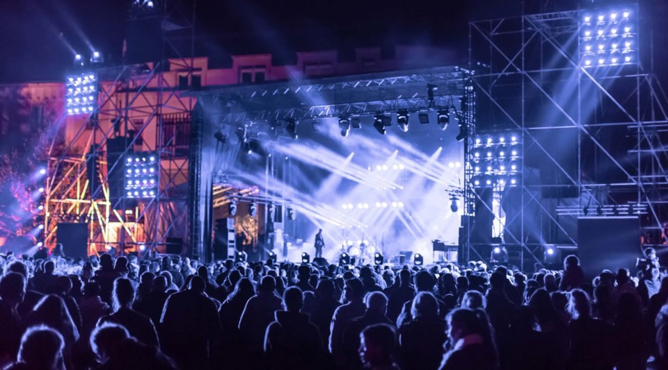 At an outdoor concert, a large crowd is gathered and the stage is illuminated with a series of bright lights and colorful effects. Powerful beams and spotlights are used to enhance the atmosphere and create a lively and energetic environment for the audience.