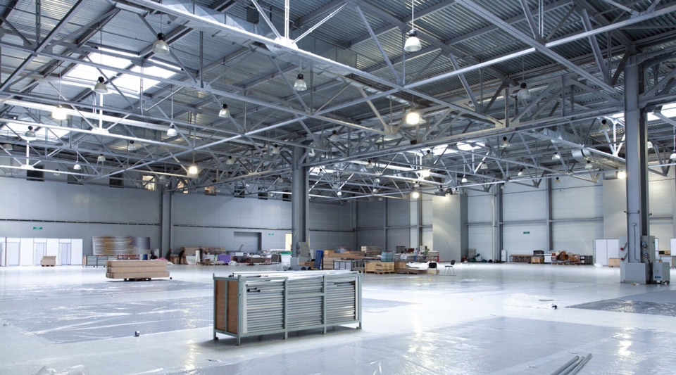 A large industrial warehouse is being prepared, with various materials and equipment being organized throughout the open space. The high ceilings and exposed beams are being illuminated by overhead lights, providing a bright and functional environment for work or storage.