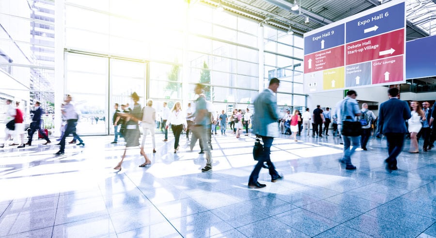 People are being seen walking through a bright and spacious convention center hallway, illuminated by natural light from large windows. Directional signs are being displayed prominently overhead to guide visitors to different expo halls and event areas.