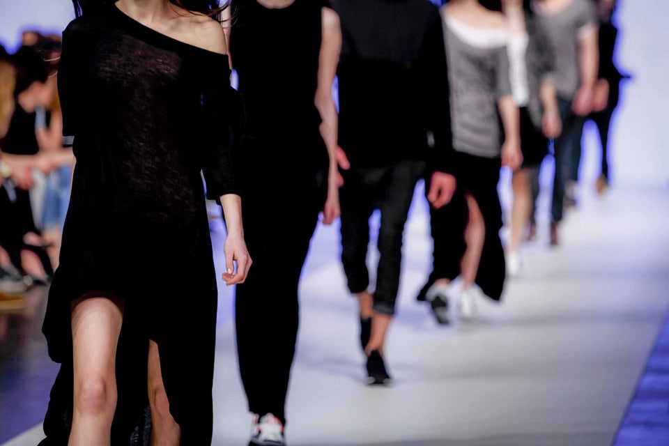 Models are being showcased walking down a runway, dressed in a variety of contemporary outfits. The fashion show is being observed by an audience seated along the sides, highlighting the latest trends in clothing design.