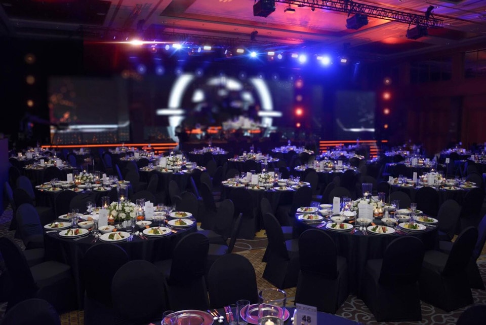 A large banquet hall is filled with numerous round tables decorated with neatly arranged plates, glasses and bouquets of flowers. The stage is illuminated with colorful lights to create a festive atmosphere for the event.