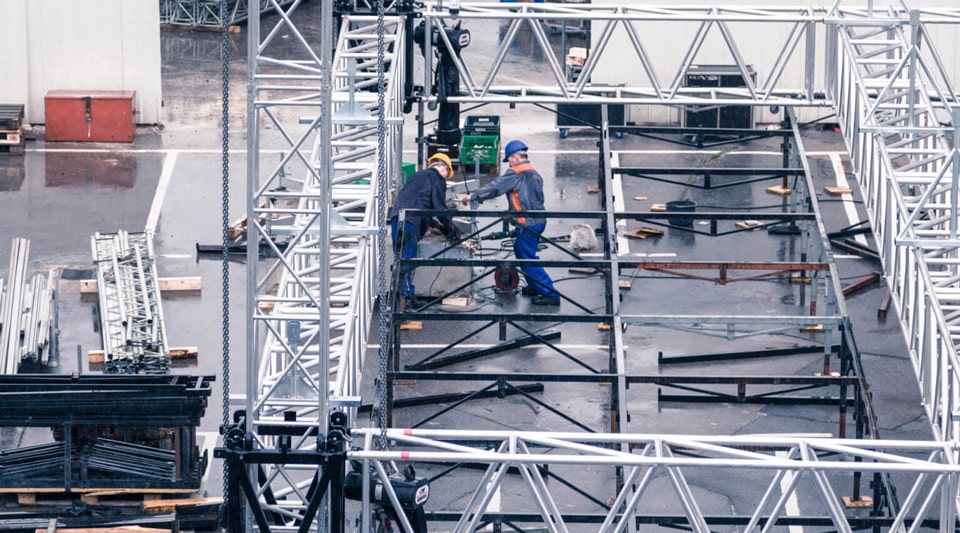 A scaffolding for a stage is being assembled by workers wearing safety helmets. Various metal parts and tools are being used to construct and secure the structure.