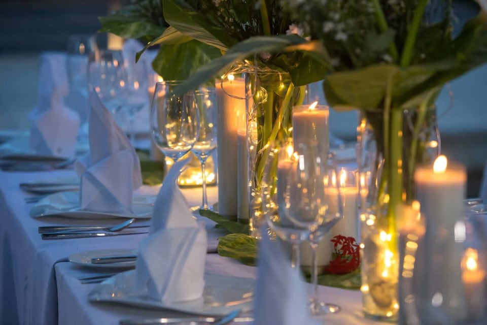 The table is elegantly set with neatly folded napkins, glasses and cutlery and decorated with floral arrangements. Soft candlelight creates a warm and intimate atmosphere for dinner.