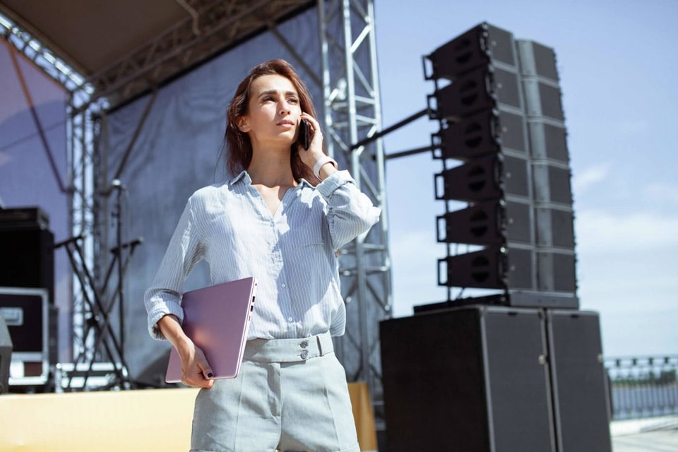 A woman is being shown at an event, talking on the phone and holding a laptop. The stage in the background is being prepared with professional equipment for a presentation.