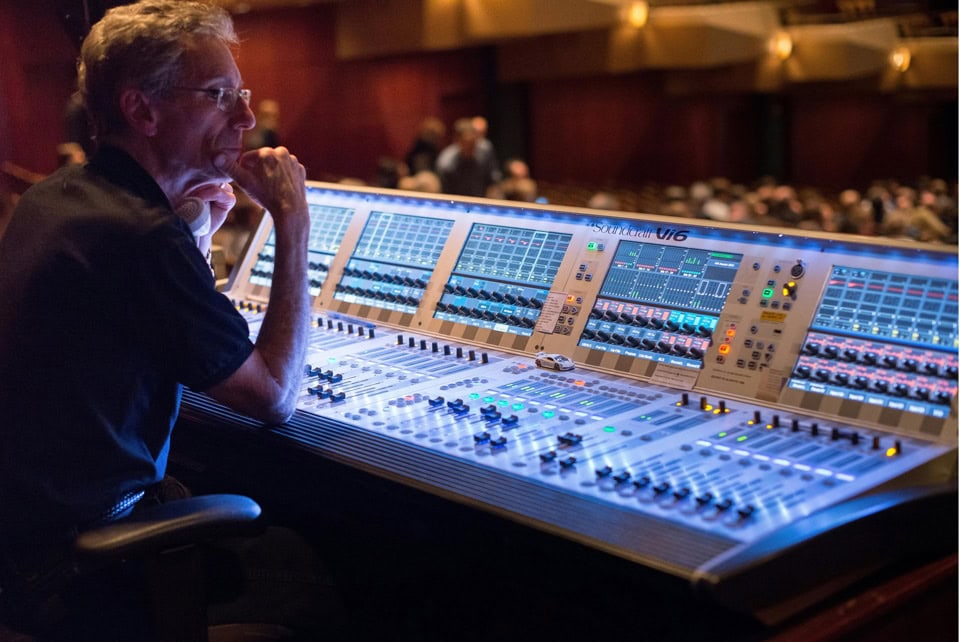 A sound technician is being shown working at a mixing console to control the audio quality for an event. Various knobs and sliders are being used to adjust sound levels and effects.