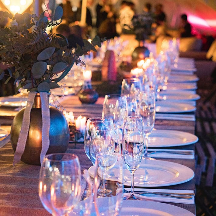 A long table is being set for an elegant event, with dishes, glasses, and candles creating a warm ambiance. Floral arrangements are being used as table decorations to enhance the festive atmosphere.