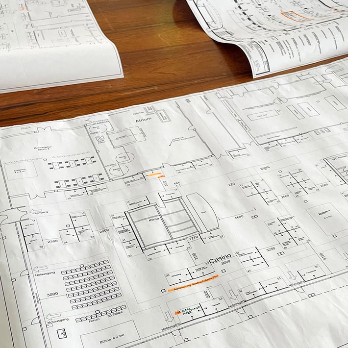 Architectural floor plans are being laid out on a wooden table, showing detailed layouts of a building's interior spaces. Specific areas and rooms are being marked with annotations and highlighted sections for emphasis or modifications.