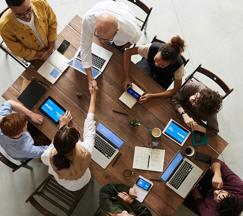 A team of people is being shown around a table, while a handshake is being exchanged between two individuals. Laptops and notebooks are being used to take notes and share information.