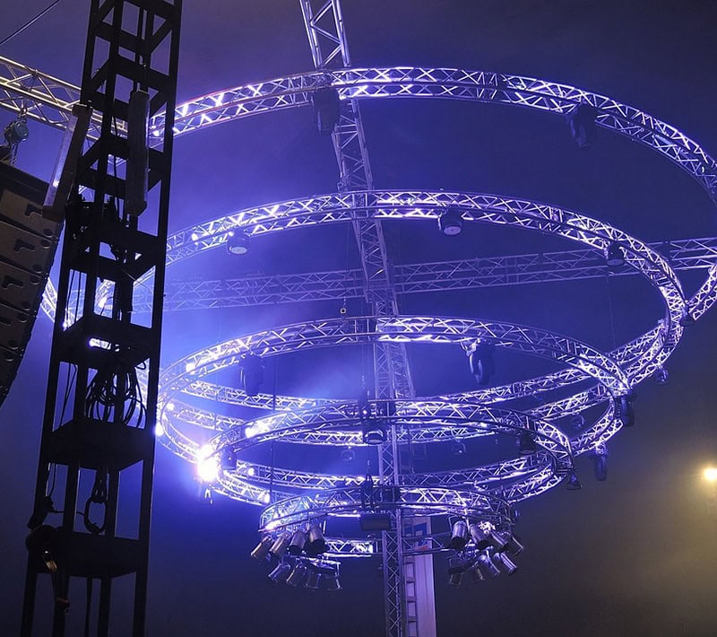 A circular lighting rig is being set up with multiple spotlights and floodlights for an event. The metal rings are being illuminated with blue lights to create an impressive visual effect.